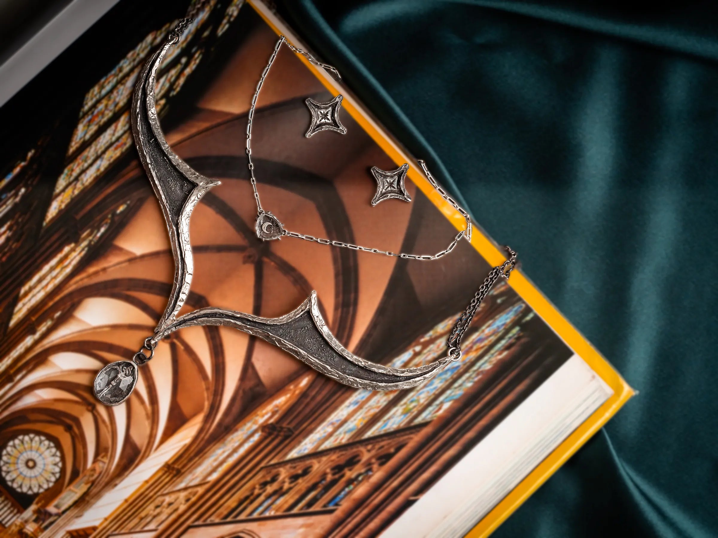 A silver geometric necklace and earrings set lies on an open book, showcasing a cathedral interior. The scene is elegant with a dark fabric backdrop.
