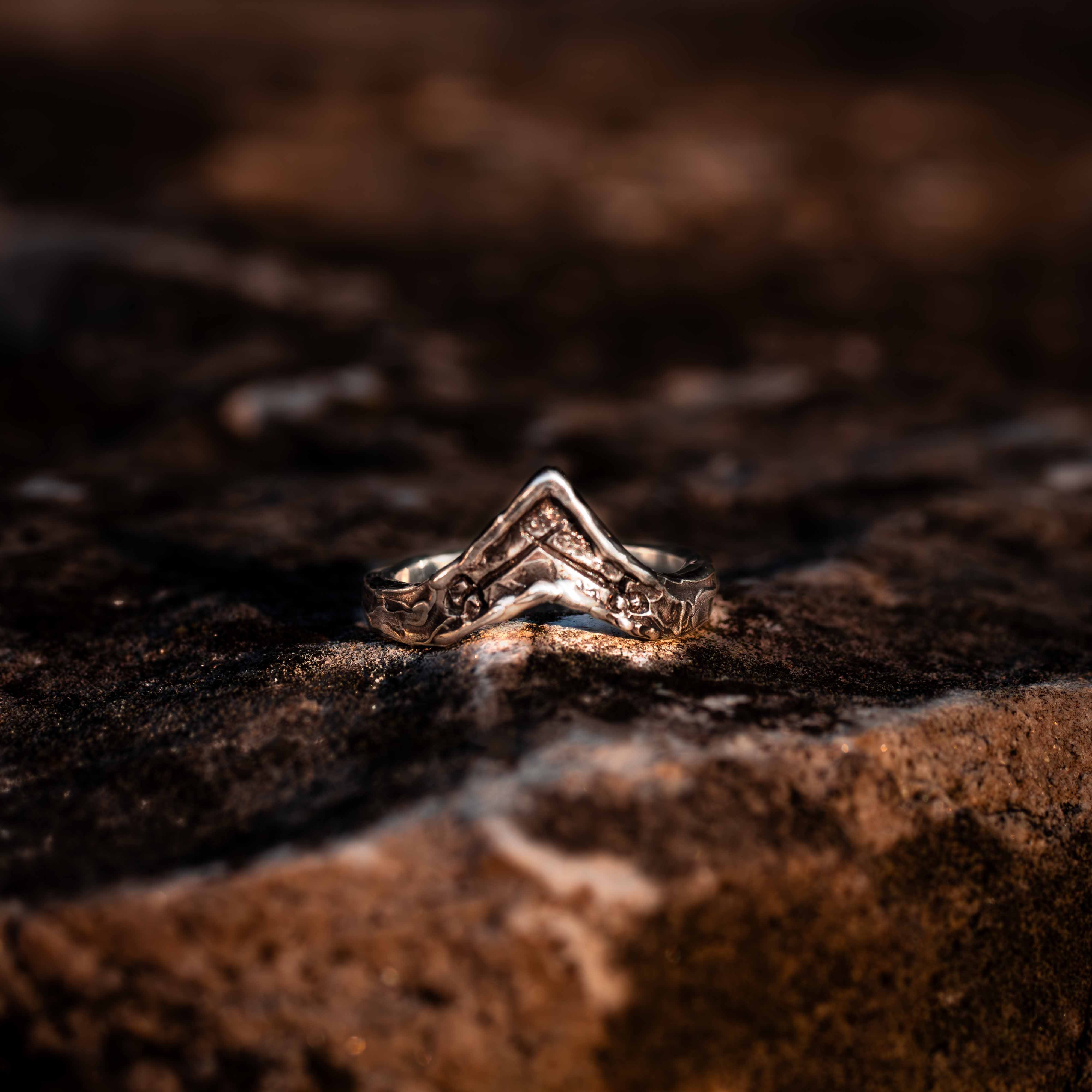 A silver ring with V-shaped shadow band and crossed sword engravings, placed on a textured dark background.