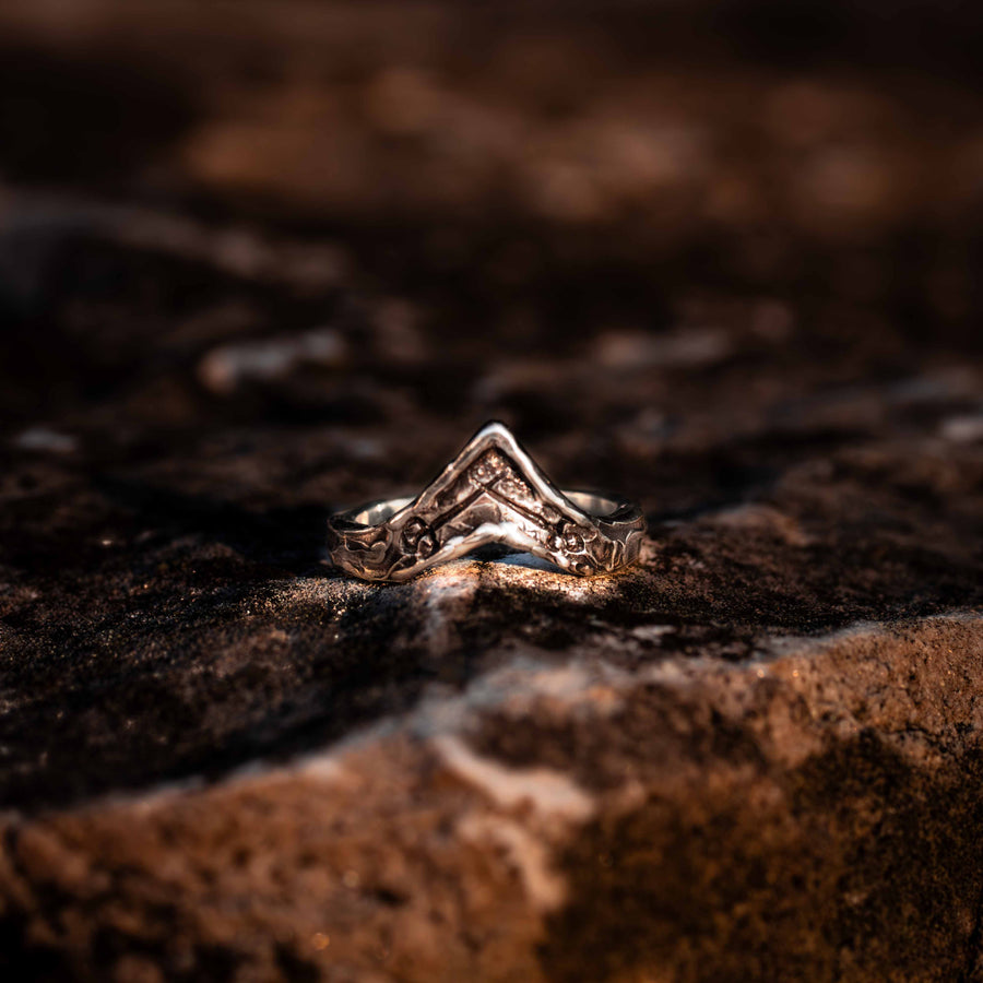 A silver ring with V-shaped shadow band and crossed sword engravings, placed on a textured dark background.