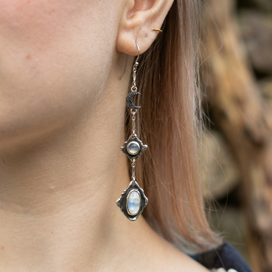 A close-up of a person's ear wearing a long silver dangle earring with a crescent moon charm, a round moonstone, and a larger faceted moonstone at the bottom.