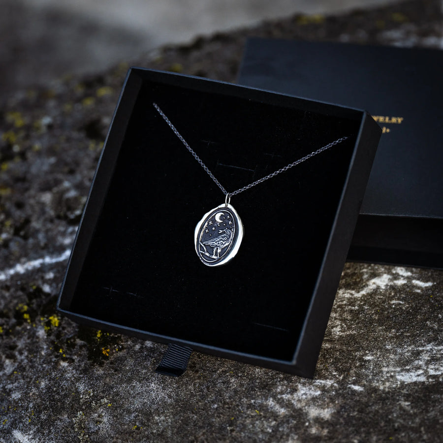 a silver necklace depicting a raven and crescent moon with wax seal styling in a black gift box