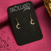 A pair of dangly gold moon earrings on a black Skolland Jewelry earring card.