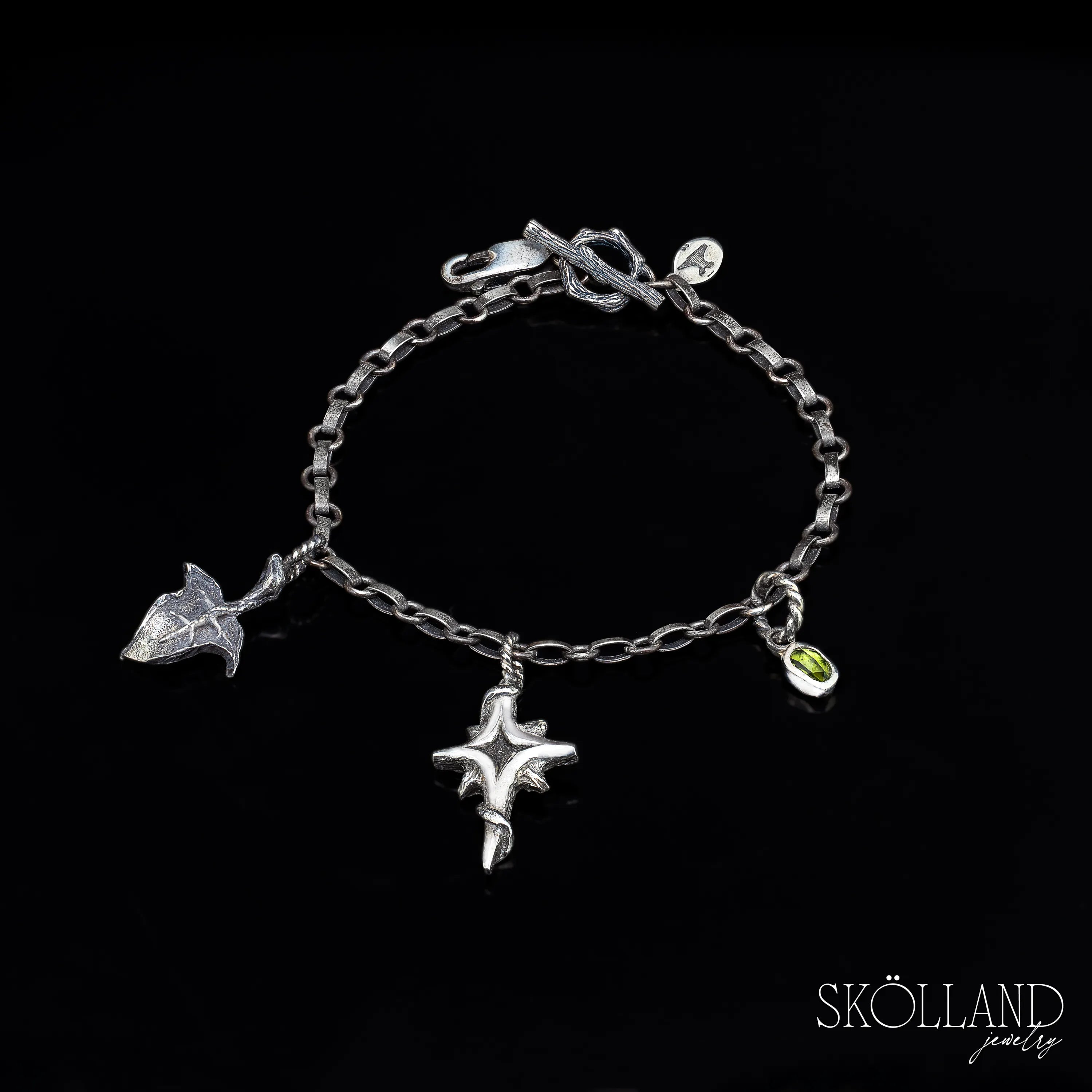 A sterling silver charm bracelet with a chain link design, featuring a charm that resembles a north star and another that appears to be a small ivy leaf with intricate detailing,and a green gemstone- tourmaline.