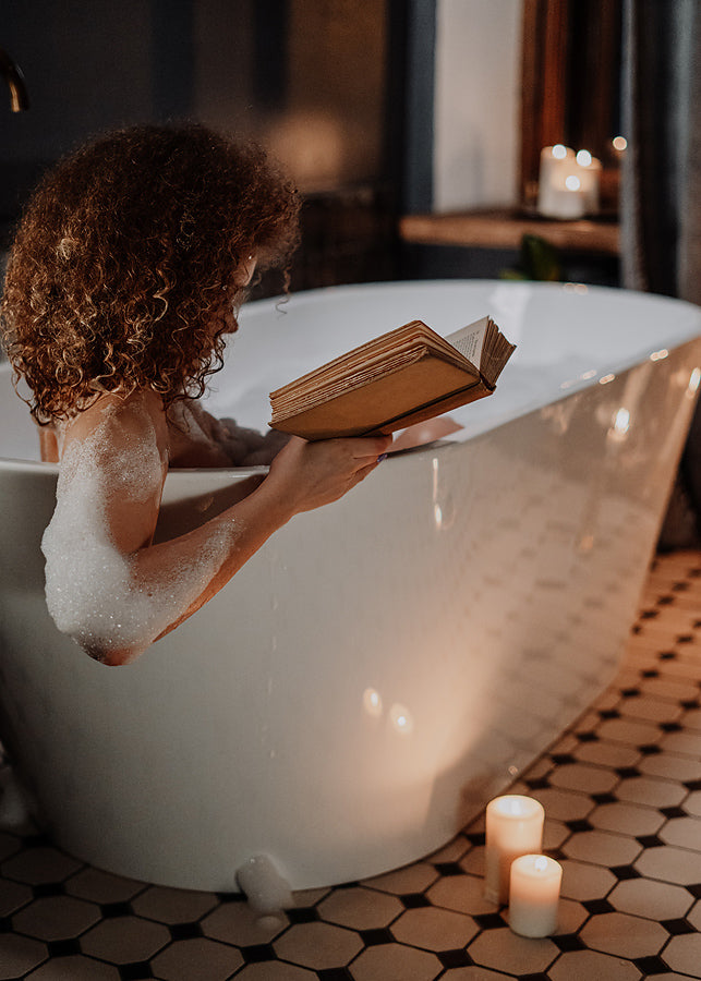 Woman reading a hard cover book in a freestanding bathtub by candlelight