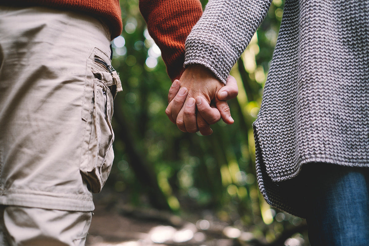Two people holding hands, one in a gray knit sweater and jeans, the other in a rust sweater and beige pants, standing in a lush forest.