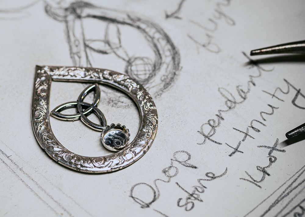Elegant silver teardrop pendant with intricate floral engravings and Celtic trinity knot design, placed on a sketch with jewelry notes.