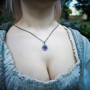 A close-up lifestyle shot of a woman wearing a silver chain with a round purple amethyst pendant, styled with a light blue ruffled dress against a blurred outdoor background.