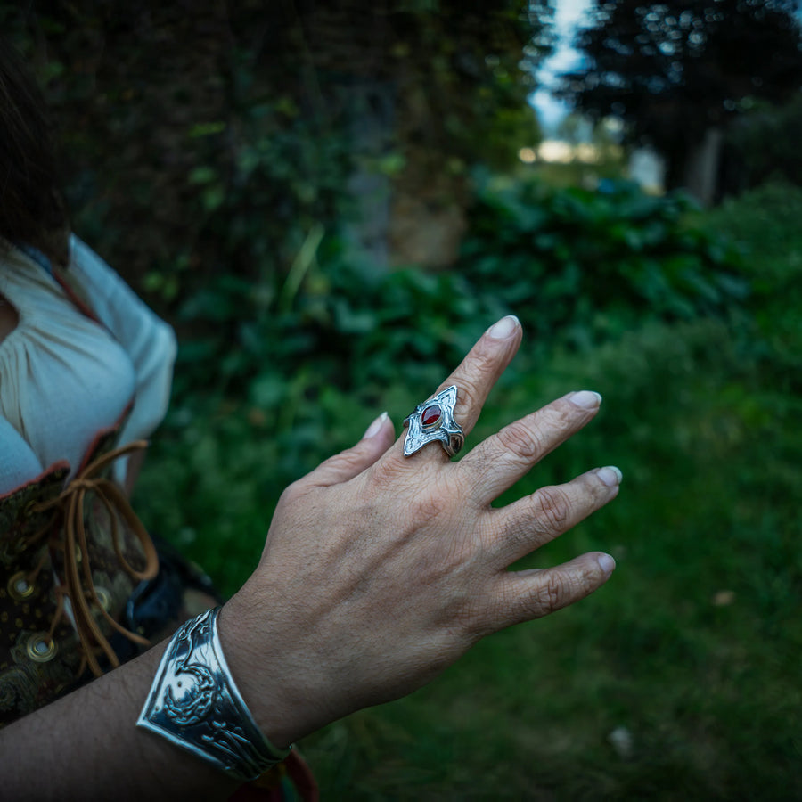 A close-up of a person's hand wearing a sterling silver ring with a red garnet gemstone and a matching silver cuff bracelet, set against a blurred outdoor background of green foliage.