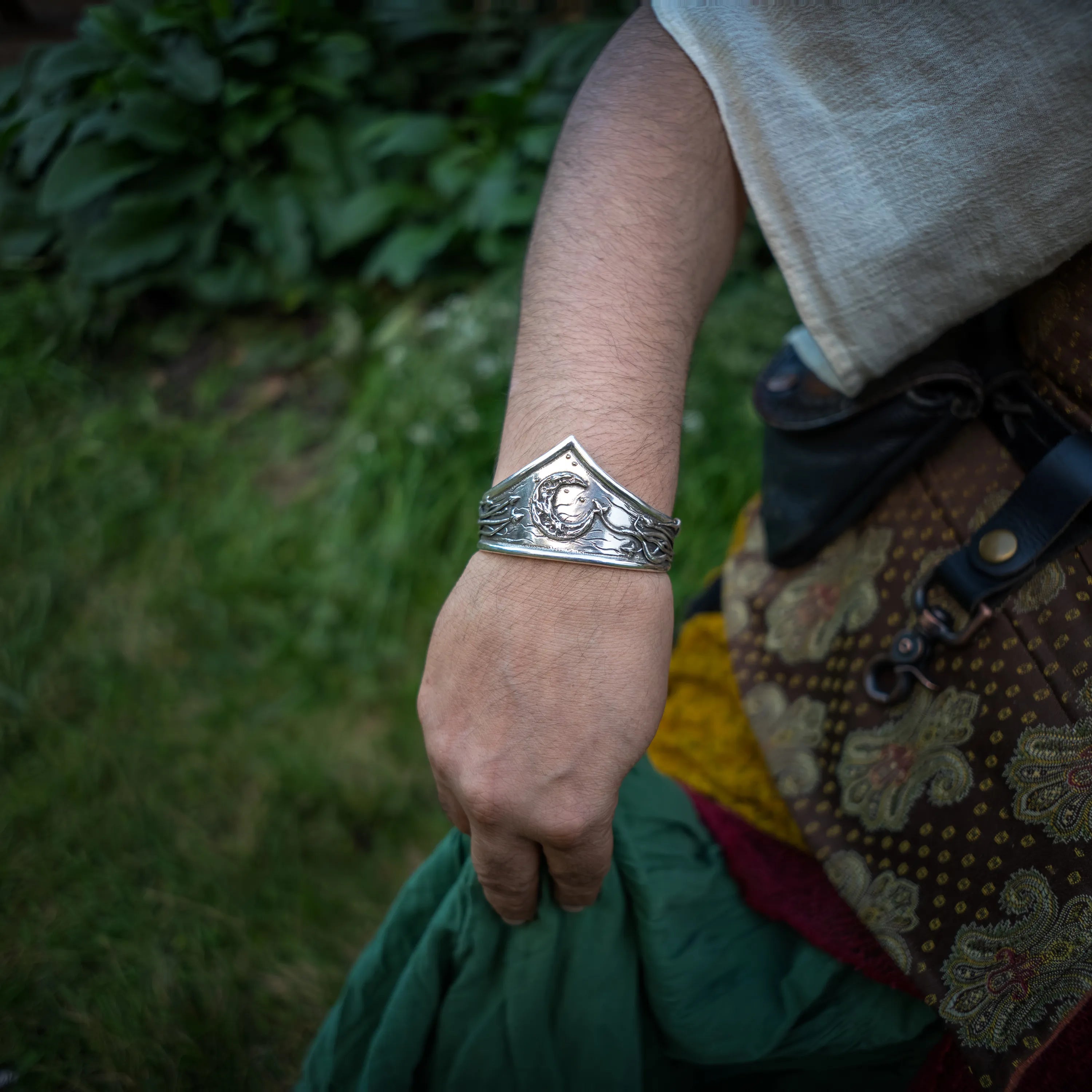 A sterling silver celestial metal cuff bracelet worn on a person's wrist, shown outdoors with grass and foliage in the background and a patterned garment visible.