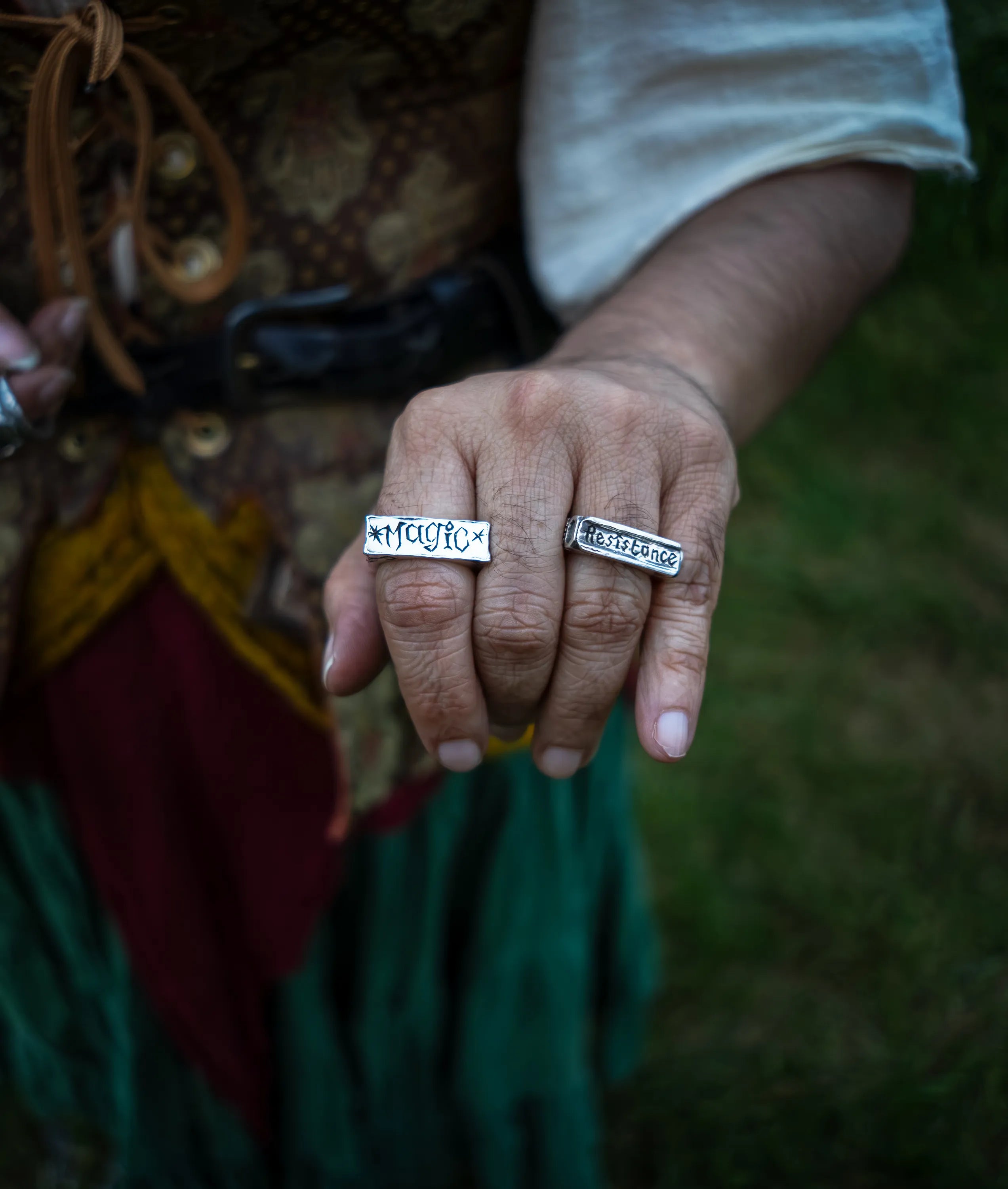 A close-up of a person's hand wearing two sterling silver rectangular signet rings with engraved text, one reads "magic" the other reads "resistance", set against a blurred outdoor background.
