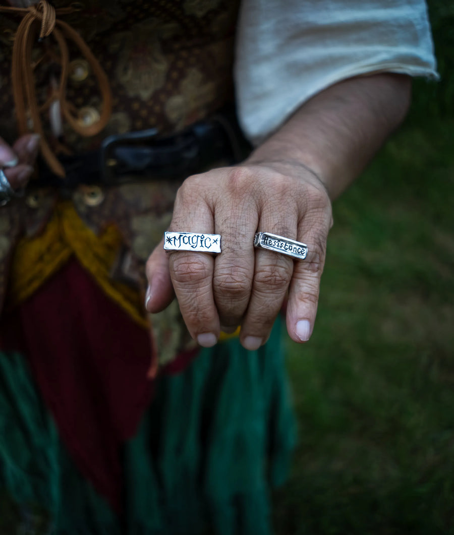 A close-up of a person's hand wearing two sterling silver rectangular signet rings with engraved text, one reads "magic" the other reads "resistance", set against a blurred outdoor background.