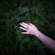 A person's hand resting in dark green foliage, wearing a statement argentium silver ring with a starry design at the top of the ring. Moonstone is set in the center of the star.