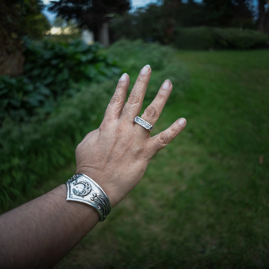 A person's hand and wrist wearing a sterling silver cuff bracelet and a signet ring that reads "resistance", set against a blurred green garden background.