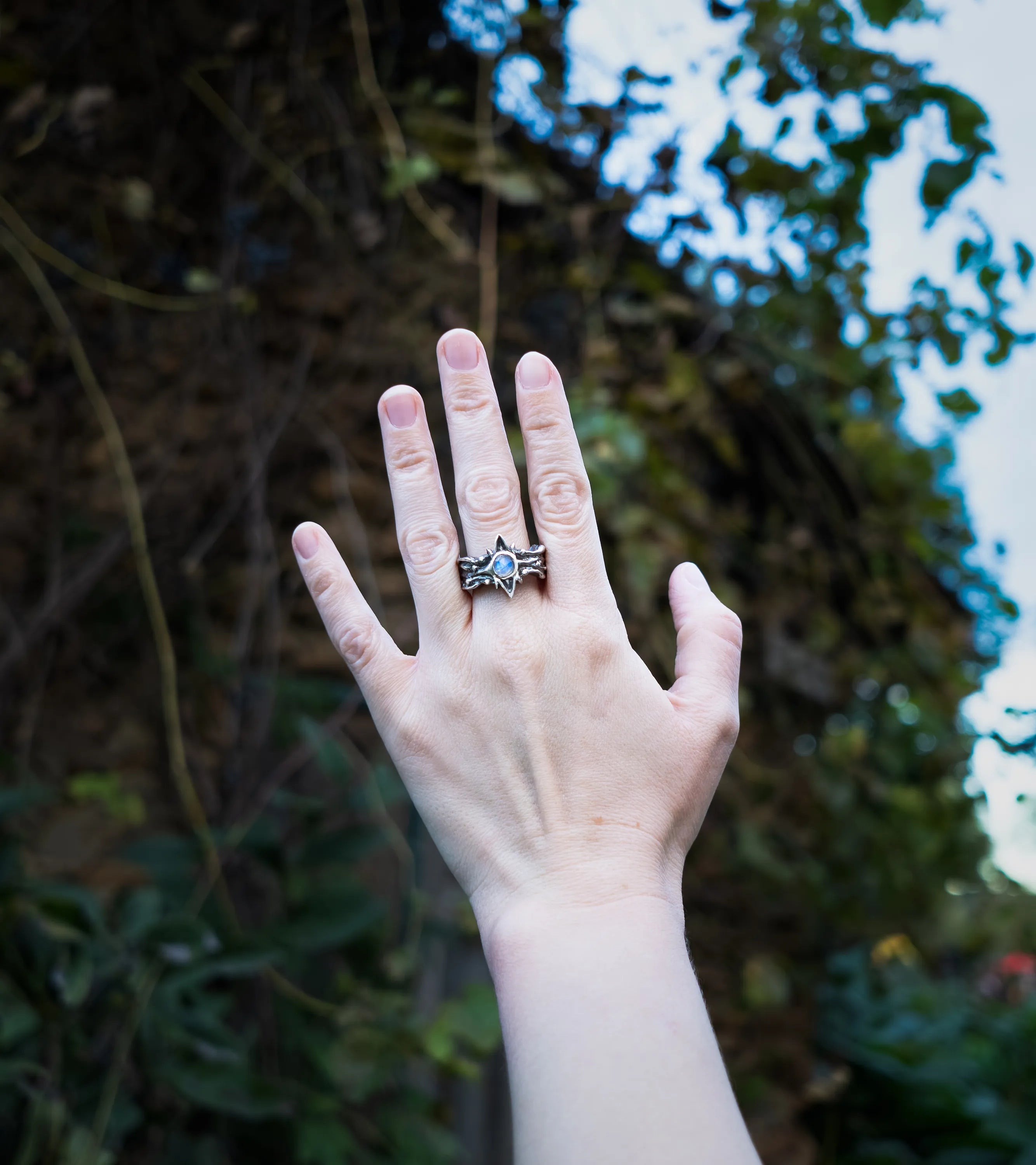 A hand wearing a sterling silver ring with a blue moonstone inside of a star center, set against a blurred outdoor background of green foliage and sky.