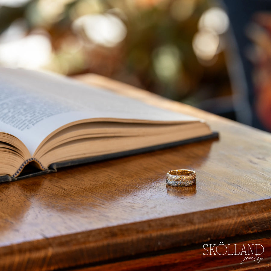 Gold rings on a wooden surface next to an open book with a blurred background. One gold ring has a dark black rhodium finish.