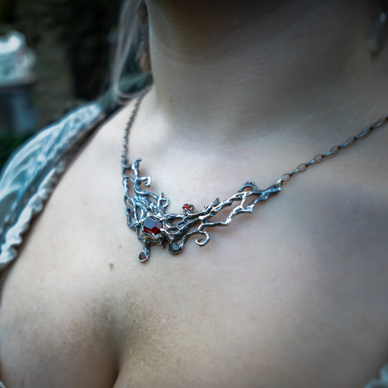 A close-up lifestyle shot of a person wearing a detailed silver necklace with a vine design and small red garnet accents.