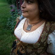 A woman wearing a brown and gold patterned medieval-style vest over a cream blouse, wearing a silver gothic pendant necklace that features pentacle and celestial motifs, photographed outdoors in a garden setting.