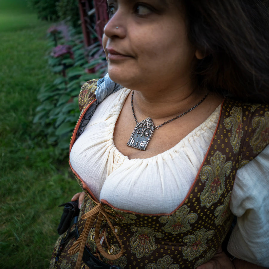 A woman wearing a brown and gold patterned medieval-style vest over a cream blouse, wearing a silver gothic pendant necklace that features pentacle and celestial motifs, photographed outdoors in a garden setting.