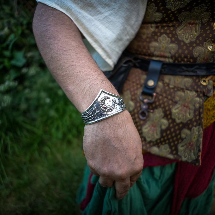 A close-up of a person's hand wearing a wide silver cuff bracelet with an embossed celestial and ivy vine design, set against a blurred outdoor background.