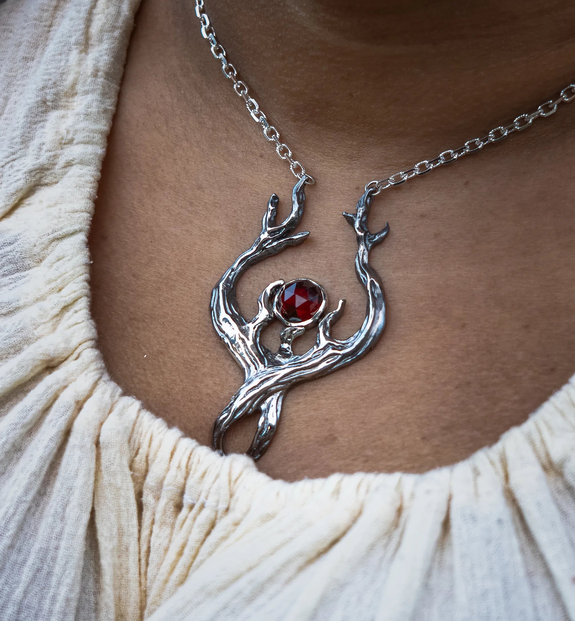A close-up of a person wearing a silver necklace with a stylized tree or flame design featuring a faceted red garnet at the center, styled with a cream-colored textured top.