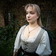 A woman wearing a medieval-style costume dress with a patterned corset bodice, white puffed sleeves, a dark green sash, and a bold,spikey v shaped necklace that is made of both sterling silver and gold, standing outdoors in front of a rustic stone wall and greenery.