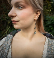 A close-up side profile of a woman wearing a long, ornate sterling silver crescent moon dangle earring with moonstone accents, set against a blurred outdoor background of green foliage.