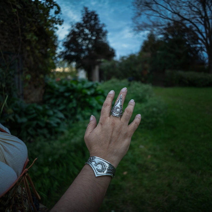 A person's hand and forearm are shown outdoors wearing a sterling silver embossed ring featuring a crescent moon on the index finger and a matching silver-cuff bracelet, with a blurred garden background.