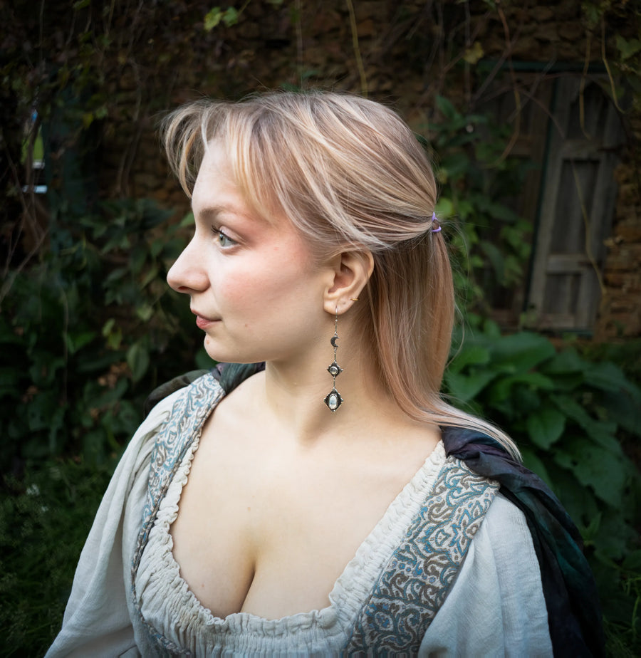 A woman in profile wearing a medieval-style dress wearing dangling moonstone earrings that feature a crescent moon, shown outdoors against a background of green foliage and a stone wall.