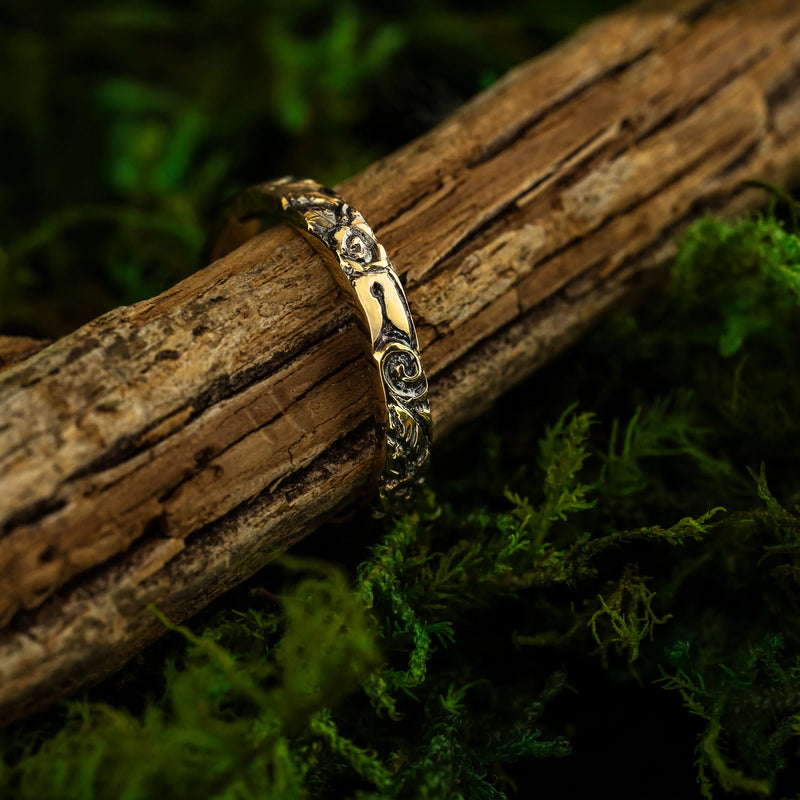 A 14K yellow gold band with hand-carved floral designs including swirls, roses, and leaves, displayed on a wooden surface with a green mossy background.