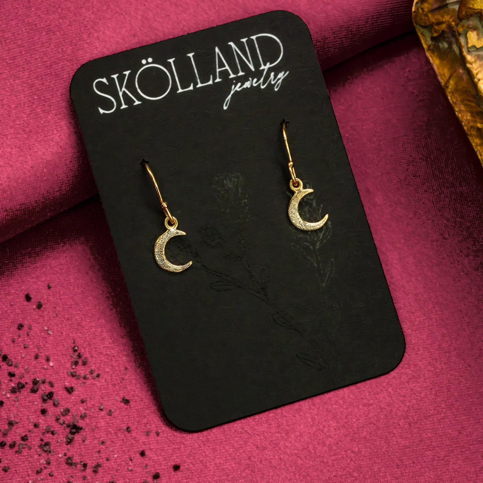 A pair of dangly gold moon earrings on a black Skolland Jewelry earring card. 