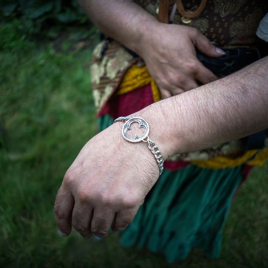 A close-up of a person's wrist wearing a sterling silver chain bracelet with a circular gothic trefoil design, set against a blurred outdoor background of green grass and colorful clothing.