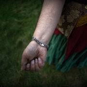 A close-up of a person's wrist wearing a sterling silver chain bracelet with a lobster clasp, set against a blurred outdoor background of green grass and patterned clothing.