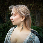 A woman in profile wearing a long silver moonstone dangle earring with dark blue-green teardrop-shaped stones, set against a blurred outdoor background of green foliage and a stone wall.