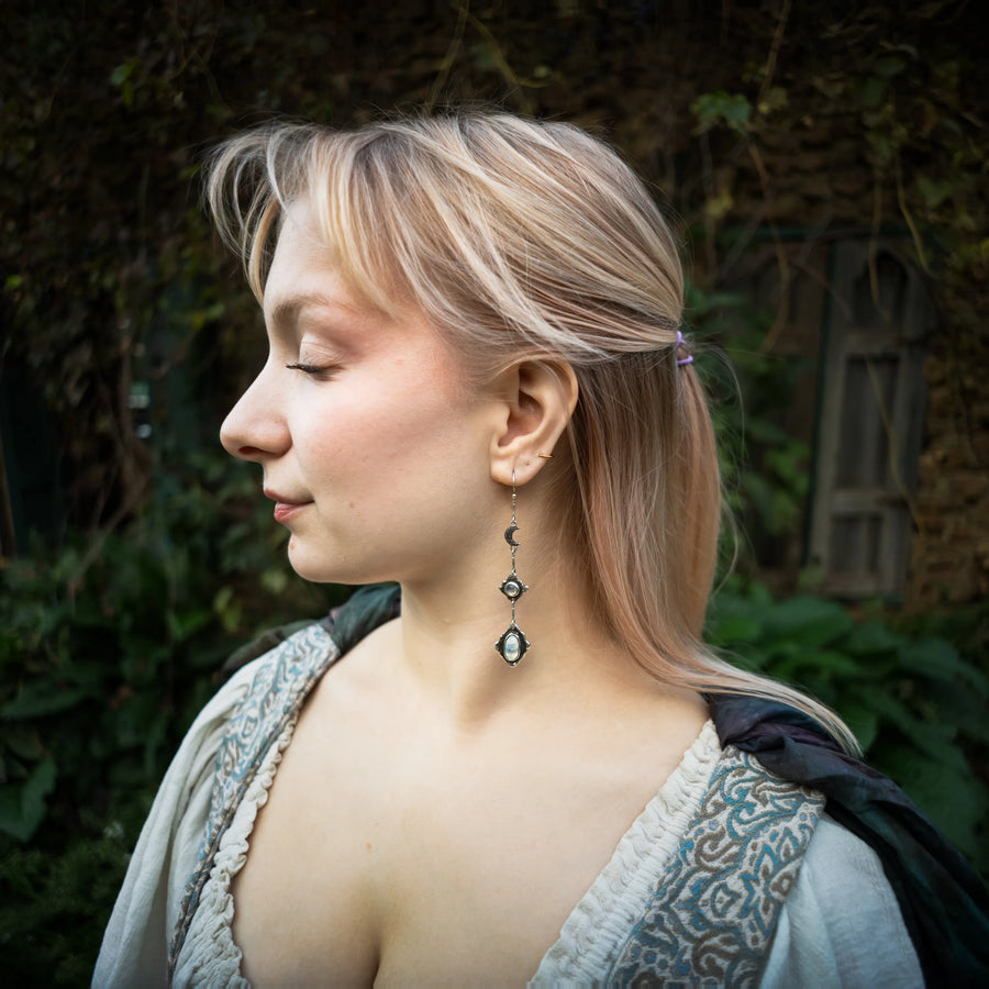 A woman in profile wearing a long silver moonstone dangle earring with dark blue-green teardrop-shaped stones, set against a blurred outdoor background of green foliage and a stone wall.