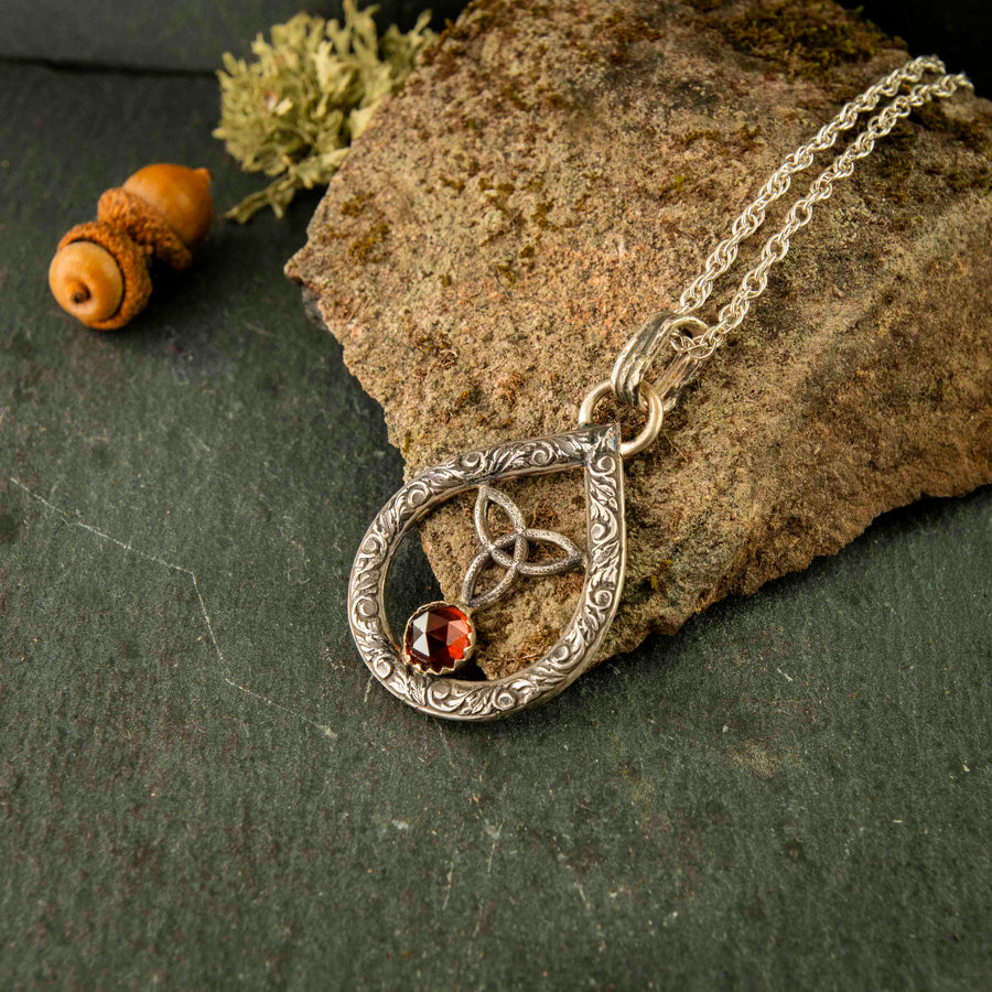 The maiden necklace, a tear dropped shaped sterling silver pendant that freatures a trinity knot, rosecut garnet and filigree styling. Handmade in the USA with ethical craftsmanship