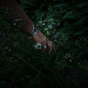 A close-up of a person's hand wearing a wide silver cuff bracelet and a blue moonstone ring, gently touching small white flowers amidst dark green foliage in a dim outdoor setting.