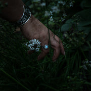 A close-up of a hand reaching toward small white flowers in a dark garden, wearing a sterling silver ring with a light blue moonstone and a silver bracelet.