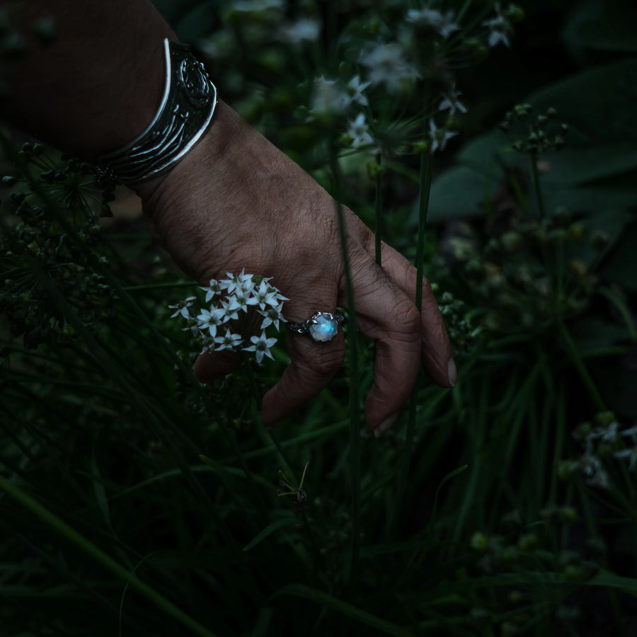 A close-up of a hand reaching toward small white flowers in a dark garden, wearing a sterling silver ring with a light blue moonstone and a silver bracelet.