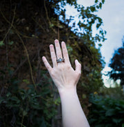 A hand wearing a sterling silver ring with a star-shaped setting and moonstone set in the center, photographed outdoors against a blurred background of foliage and sky.