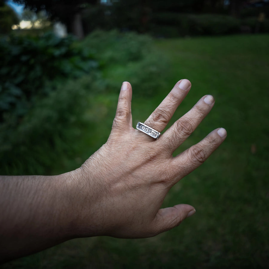 A sterling silver rectangular signet ring engraved with 'RESISTANCE' is worn on a person's ring finger, shown against a blurred green outdoor background.