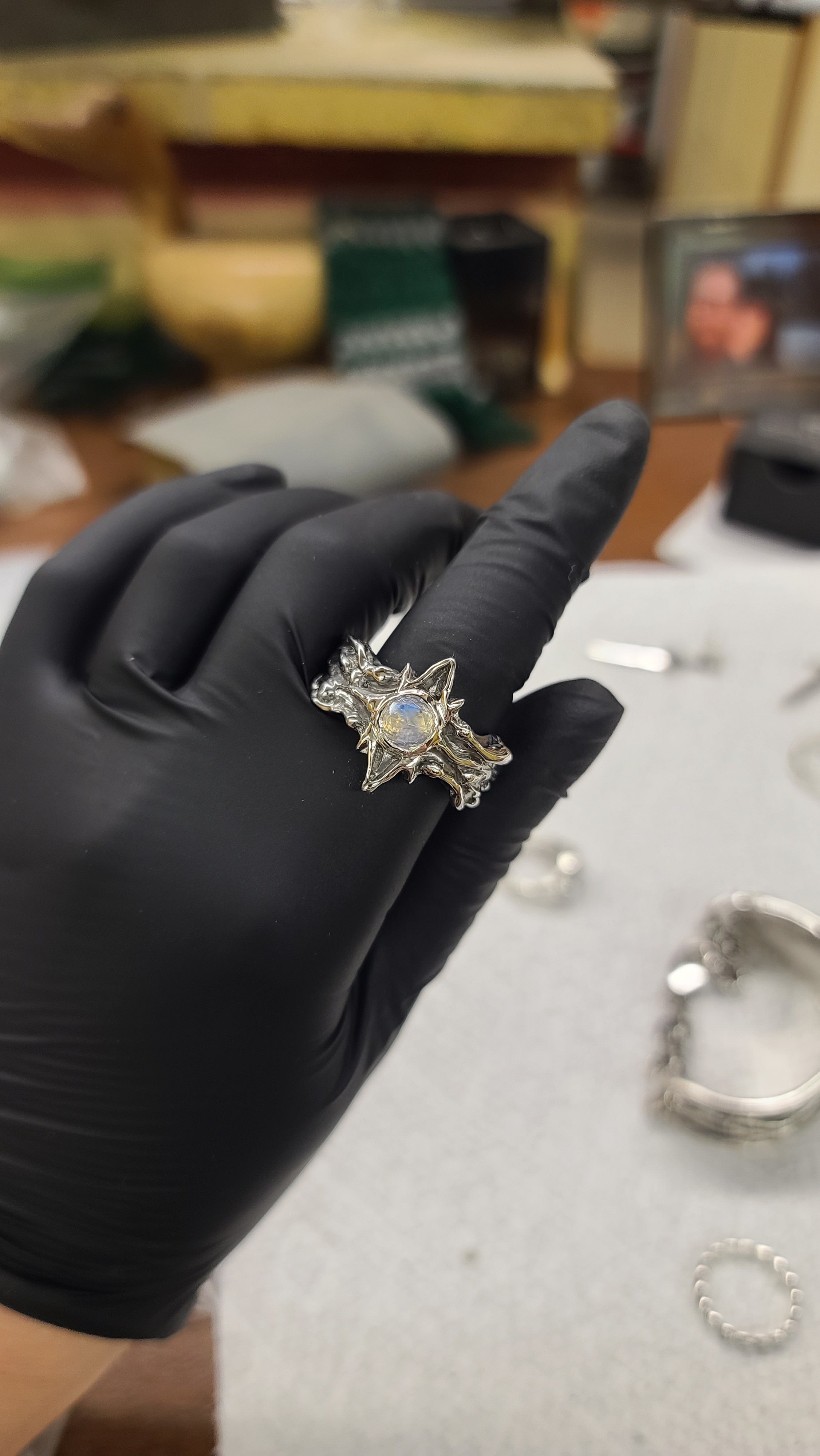 A sterling silver star ring with a light blue moonstone, held on a finger wearing a black glove in a workshop setting.