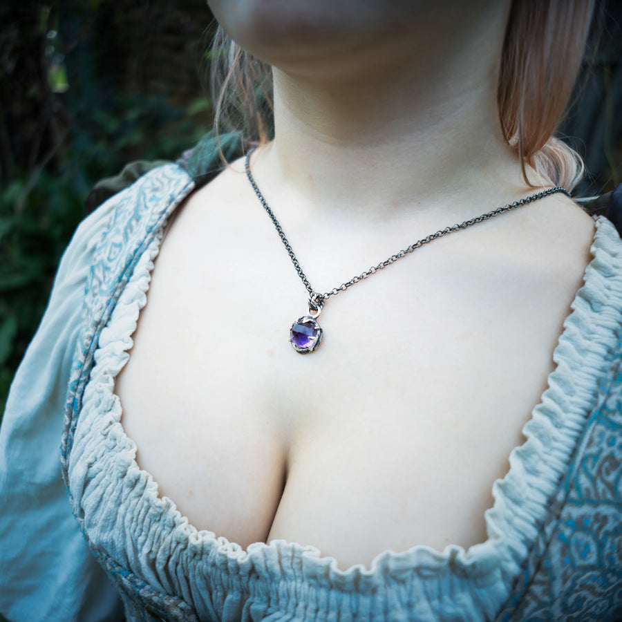 A close-up lifestyle shot of a woman wearing a sterling silver chain with a round purple amethyst pendant, styled with a light blue ruffled dress against a blurred outdoor background.