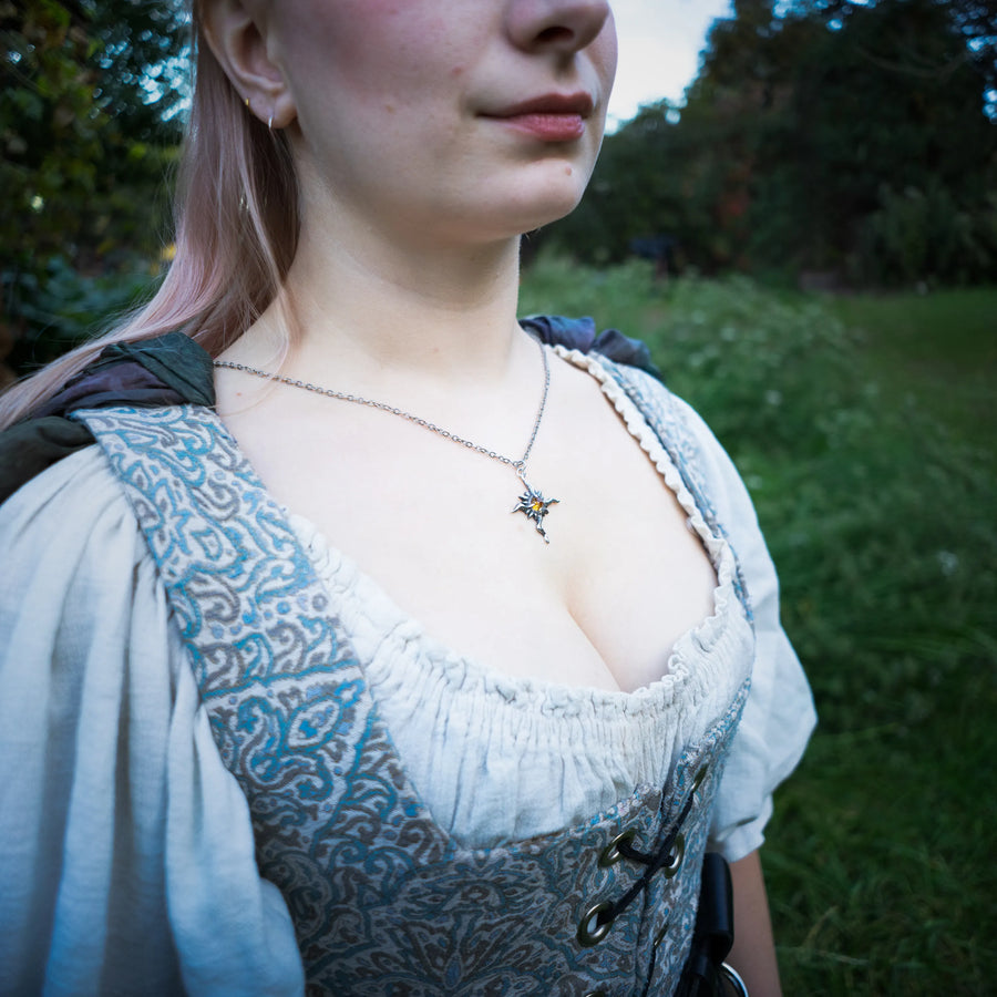 A close-up lifestyle shot of a woman outdoors wearing a medieval-style blue and white patterned corset dress over a white blouse, accessorized with a sterling silver chain featuring a golden citrine sun pendant.