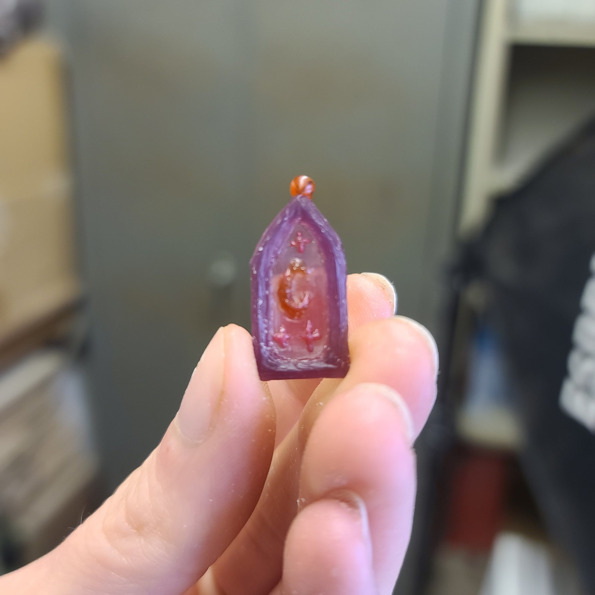 Hand holding the original wax version of this pendant before it was cast. The wax features 3 stars, a moon, and a hand sculpted gothic style border.