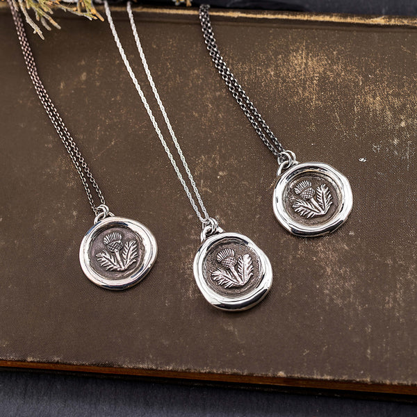 Three sterling silver necklaces with wax seal thistle pendants laid out on an old book. Each necklace has a different chain styles and the pendants feature an antiqued thistle design.
