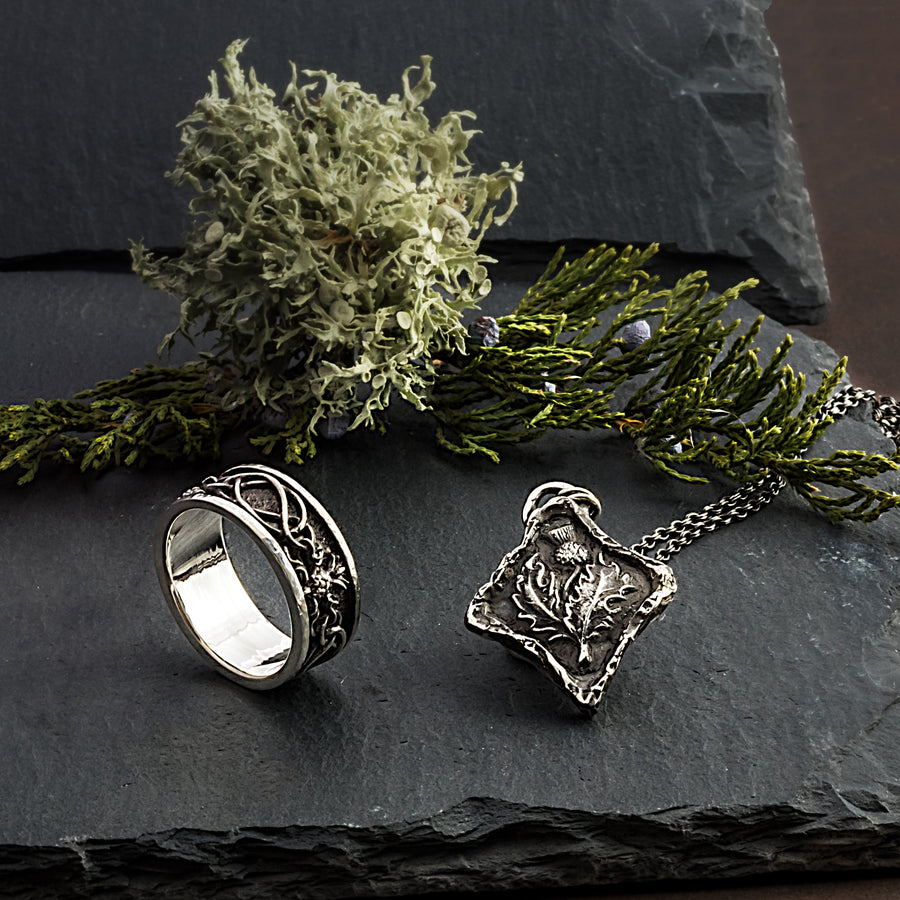 A photo of the matching thistle & highland interlace ring with the diamond shape thistle necklace that this listing is for. In the background are some juniper branches and moss.